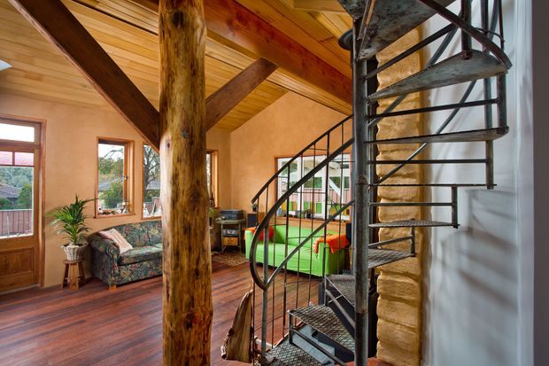 Rustic open-plan living area with a dramatic spiral metal staircase, exposed natural timber post and beams, warm timber vaulted ceiling, polished dark timber flooring, and earth-toned plaster walls. A patterned sofa, indoor plants, and colourful furnishings create a relaxed, eclectic atmosphere, with large windows providing natural light.