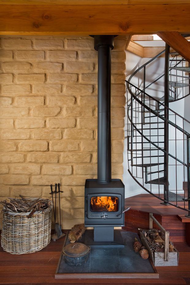 Interior view centered on a freestanding black wood-burning stove with visible flames, set against a warm textured tan brick wall. A dark timber ceiling beam spans overhead, while a black metal spiral staircase rises beside the stove to the right, with a skylight above. The room has polished dark timber flooring, a slate hearth, firewood storage, and a woven basket, creating a rustic, warm architectural interior.