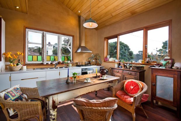 Warm, eclectic kitchen-dining space with timber-lined ceiling, earthy plaster walls, timber-framed windows, white cabinetry, and a large distressed timber dining table. Wicker armchairs, colourful cushions, plants, and stained-glass-style window panels create a relaxed, characterful interior. A compact kitchen with rangehood, cooktop, sink, and cabinetry occupies the rear of the room.