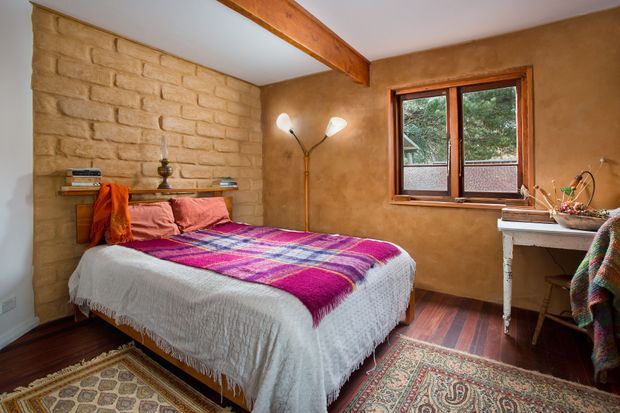 Warm, rustic bedroom with a timber-beamed ceiling, exposed light-coloured brick feature wall, earthy textured plaster walls, dark timber flooring, and a double bed layered with colourful woven textiles. A timber-framed window overlooks greenery, while a small writing desk and vintage-style floor lamp complete the room.