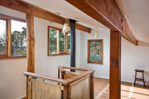 Interior view of a rustic timber-framed upper-level landing or open-plan living area. Exposed rough-hewn wooden beams and posts structure the room, with warm timber-framed windows, a black cylindrical fireplace flue, pendant lights, pale walls, and a timber balustrade around the stair opening. Trees are visible through the windows.