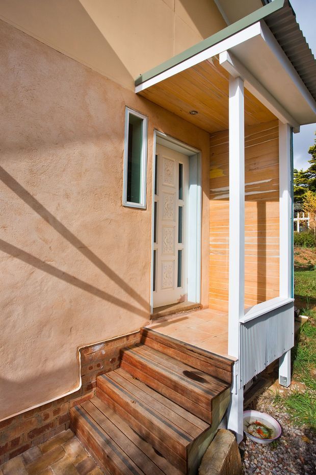 Exterior view of a compact covered front entry porch with timber steps, a decorative pale front door, rendered walls, timber-lined porch ceiling and wall, white structural posts, and a corrugated roof edge. The entry is raised slightly above a landscaped gravel and garden area.