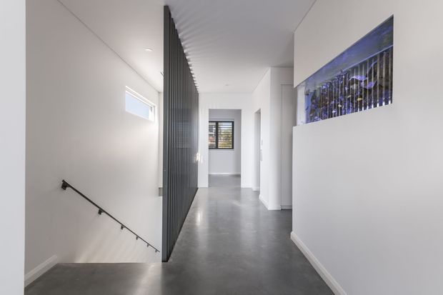 A bright, minimalist interior hallway or landing with polished grey concrete flooring, white walls and ceiling, and a dark vertical metal screen dividing the stairwell from the circulation space. A simple black handrail descends along the stair opening, while a narrow high window and a blue-toned glazed feature opening add detail. The space has clean contemporary lines and strong natural light.