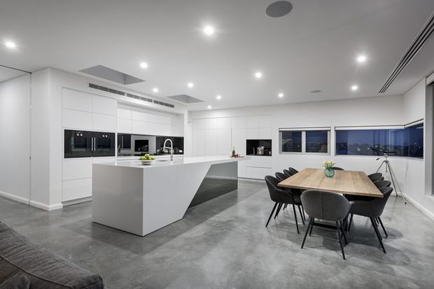A spacious open-plan contemporary kitchen and dining area with extensive white handleless cabinetry, integrated black appliances, a large white waterfall-edge island, polished concrete flooring, and a timber dining table with dark upholstered chairs. Recessed downlights, linear air vents, and large corner windows create a bright, minimalist interior with nighttime views outside.