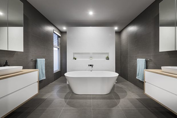 A symmetrical contemporary bathroom featuring a freestanding white bathtub, double vanity units with white cabinetry and light timber benchtops, dark grey tiled walls and floor, recessed shelving, wall mirrors, and soft blue towels. Recessed ceiling lights and a central window provide illumination.