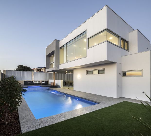 Modern two-storey residence viewed from the landscaped backyard, featuring crisp white cladding and rendered walls, expansive corner glazing, a covered outdoor entertaining area, and a rectangular illuminated swimming pool bordered by paved surfaces and artificial lawn.