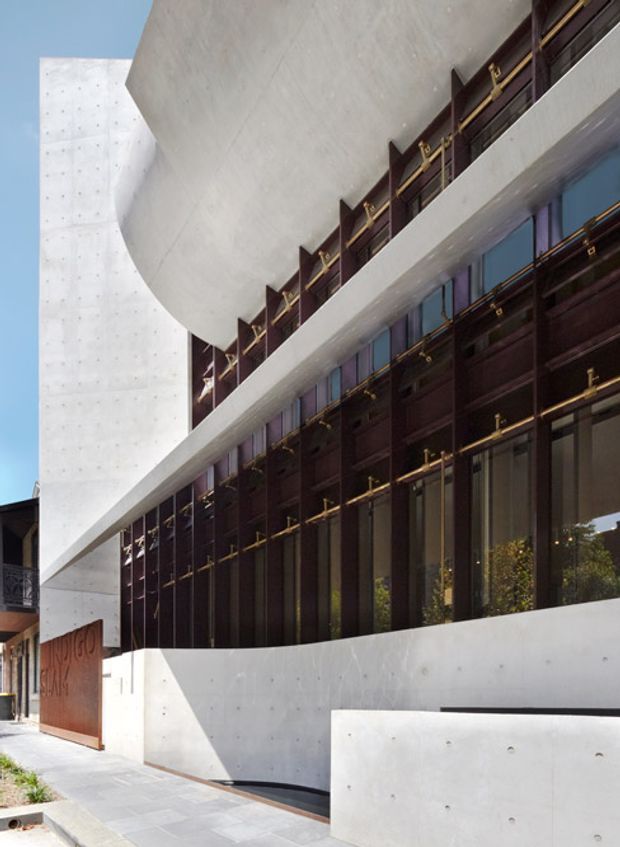 Street-level exterior view of a contemporary multi-storey building with sculptural curved and cantilevered white concrete forms, dark bronze or brown metal-framed glazing, and gold-toned horizontal and vertical detailing. A recessed entrance and paved forecourt are visible, with an adjacent white concrete wall and portions of neighbouring buildings at the edges.