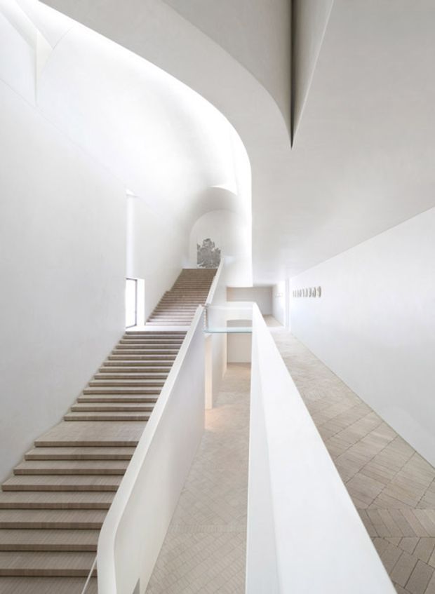 A bright, minimalist interior stairway and circulation space with broad white sculptural walls, two flights of pale stone or concrete stairs, and a long tiled ramp or corridor. Curved openings and a high, angular ceiling create a dramatic, gallery-like atmosphere, with sparse detailing and abundant diffuse daylight.