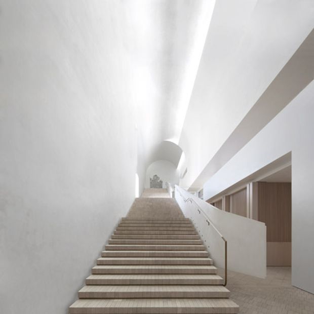 A minimalist interior stairway viewed from the bottom, with a broad flight of pale wood or stone steps ascending through a tall, bright white architectural space. Smooth sculptural walls, a curved arched opening, concealed linear lighting, and a simple white balustrade create a clean, gallery-like composition. Vertical timber panelling and a secondary corridor are visible to the right.