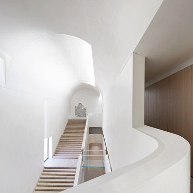 A bright, sculptural interior stairway with broad timber-toned treads rising through a tall white space. The staircase and upper landing are enclosed by smooth, curving white plaster walls and balustrades, with a clear glass guardrail and a warm vertical timber-panelled wall visible to the right. Arched openings and strong natural light create a refined, minimalist architectural composition.