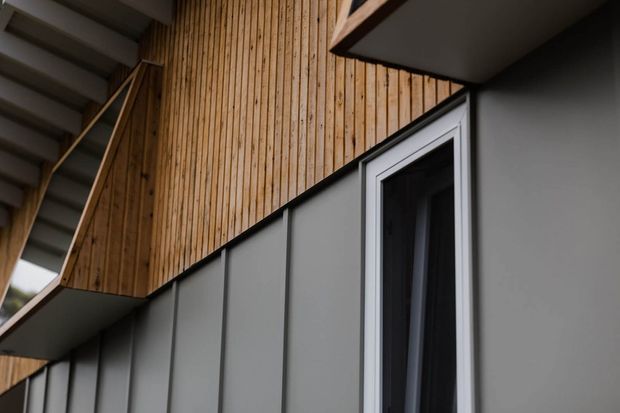 Close-up architectural exterior showing a contemporary facade with dark grey vertical metal cladding, warm vertical timber screening or cladding above, a narrow white-framed window, and strong angular roof or balcony forms. The composition emphasizes layered facade materials and geometric lines.