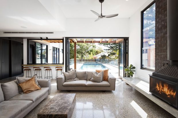Bright contemporary open-plan living room with large sliding glass doors opening to a pool terrace. The room features pale upholstered sofas, a dark kitchen and bar area with stools, polished terrazzo flooring, white walls, exposed timber pergola elements, a ceiling fan, and a built-in dark fireplace set against an exposed brick wall.