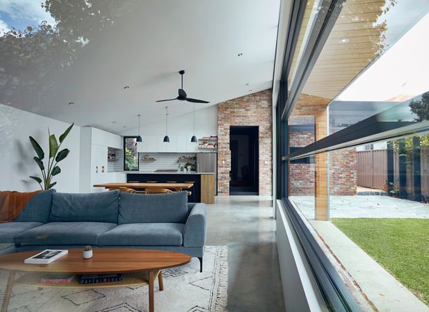 Open-plan contemporary living area with a blue upholstered sofa and timber coffee table in the foreground, flowing into a white kitchen with island bench and pendant lights. Full-height glazing provides views to a landscaped courtyard and pool. Exposed red brick walls, polished concrete flooring, timber ceiling lining, and a large ceiling fan create a warm modern interior with strong indoor-outdoor connection.