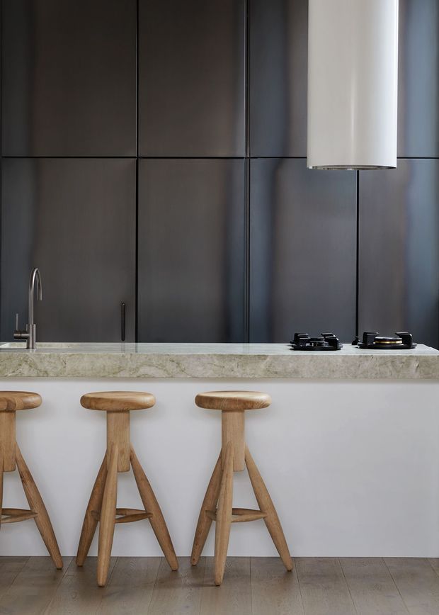 A refined contemporary kitchen with a dark, grid-panelled backsplash and integrated cabinetry, a pale stone island benchtop, and a light neutral island base. Three sculptural timber bar stools line the island, with a stainless-steel tap, black gas cooktop, and cylindrical pendant light completing the minimalist composition. The floor is a pale timber finish.