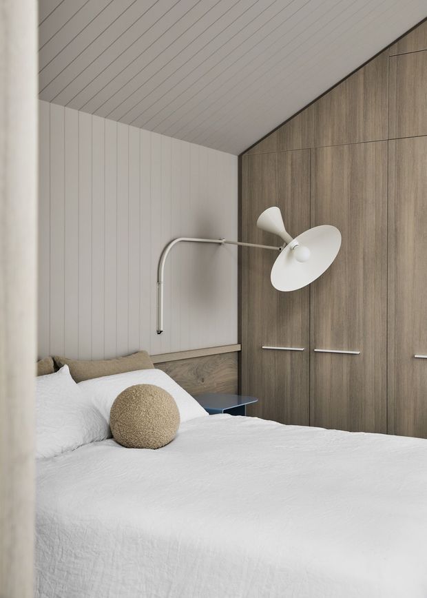 A calm, minimalist bedroom with a neatly made white bed, beige textured cushions, integrated timber wardrobe cabinetry, and a sculptural wall-mounted reading lamp. Vertical white wall panelling and matching ceiling boards create a clean, cohesive interior, while a small blue bedside table adds subtle contrast.
