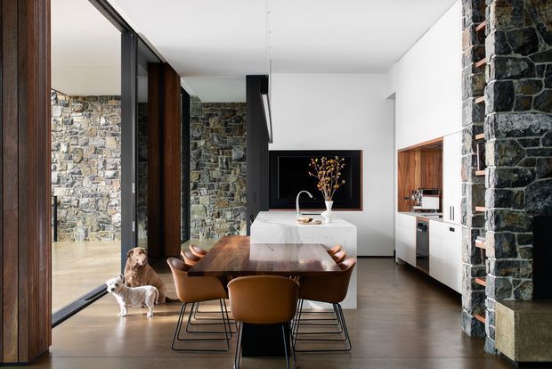 Contemporary open-plan kitchen and dining area with a long timber dining table, upholstered dining chairs, a white stone island, dark cabinetry, integrated appliances, and a large black-framed glazed opening. Rough dark stone masonry walls contrast with smooth white walls and dark flooring, while a television and decorative vase sit beyond the island. Dogs are present near the glazing.
