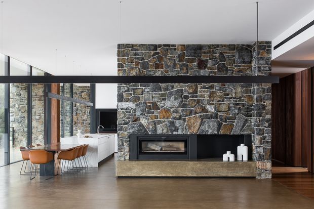 Contemporary open-plan interior dominated by a large rough natural-stone fireplace wall with a built-in firebox and long stone hearth. A dining area with a timber table and warm ochre upholstered chairs sits to the left beside extensive black-framed glazing, while a minimalist kitchen and dark timber built-in joinery are visible in the background. The room has a high ceiling, polished concrete flooring, white walls, and a restrained palette of charcoal, grey, brown, and warm timber.