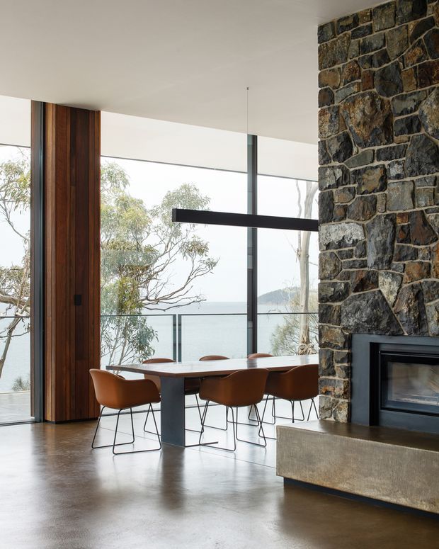 A contemporary dining area with a long timber dining table and sculptural brown chairs, set beside expansive floor-to-ceiling glazing overlooking a wooded waterfront landscape. A dramatic dark stone fireplace wall with a built-in fireplace dominates the right side, while a slim black pendant light hangs above the table. Vertical timber wall panelling and polished concrete flooring contribute to the refined, natural material palette.