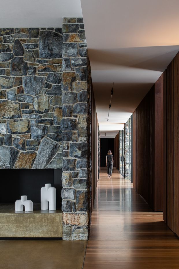 A sophisticated interior hallway with a long, narrow perspective. Irregular natural stone masonry forms a prominent structural wall and fireplace-like feature on the left, paired with dark vertical timber wall panelling and extensive built-in cabinetry on the right. Warm timber flooring, a polished concrete or stone hearth/platform, recessed linear lighting, and simple white sculptural vessels create a refined, minimalist atmosphere. A person is visible at the far end of the corridor.