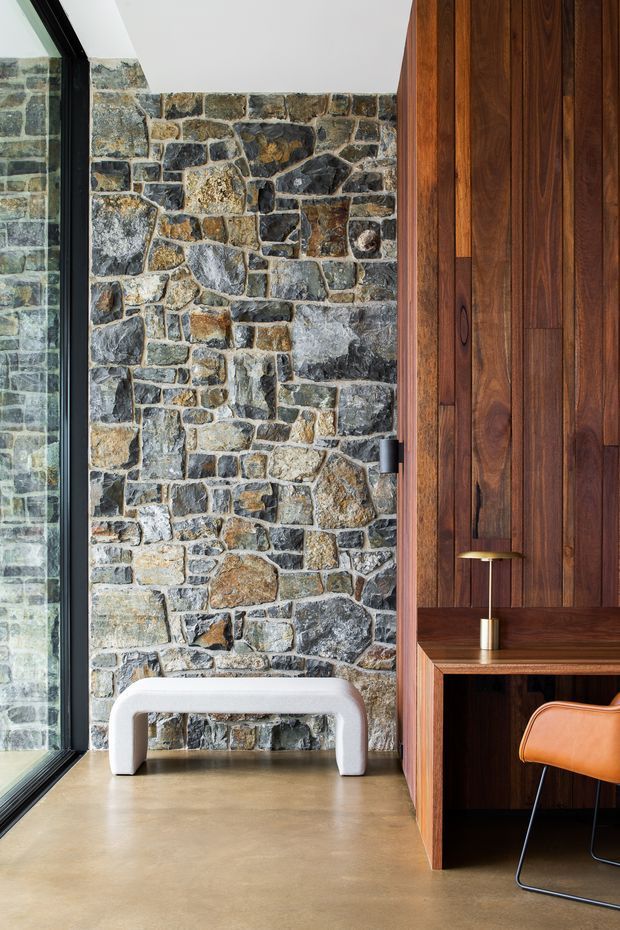 A refined interior vignette featuring a dramatic irregular natural stone masonry wall beside full-height black-framed glazing. A warm dark timber panelled built-in desk and storage unit occupies the right side, paired with a tan leather chair and small brass-toned desk lamp. A sculptural white bench sits against the stone wall, while the smooth warm-toned floor and abundant natural light create a balanced, tactile atmosphere.