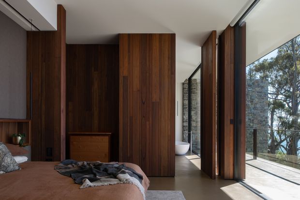 A contemporary bedroom with extensive dark timber wall panelling and built-in wardrobe joinery. The room opens toward a bright glazed corridor or balcony edge, with full-height windows and views to trees outside. A partially visible freestanding white bathtub sits beyond the bedroom, while the bed and low timber dresser define the main space. The palette is warm and earthy, with strong natural light and a refined indoor-outdoor connection.