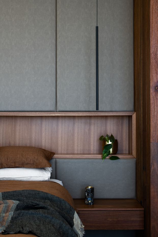 Close-up of a sophisticated bedroom wall and bed detail. The composition features dark timber wall panelling and integrated bedside cabinetry, grey upholstered panels, a slim black vertical handle, layered brown and charcoal bedding, and a small vase with greenery. The overall aesthetic is warm, moody, refined, and contemporary.