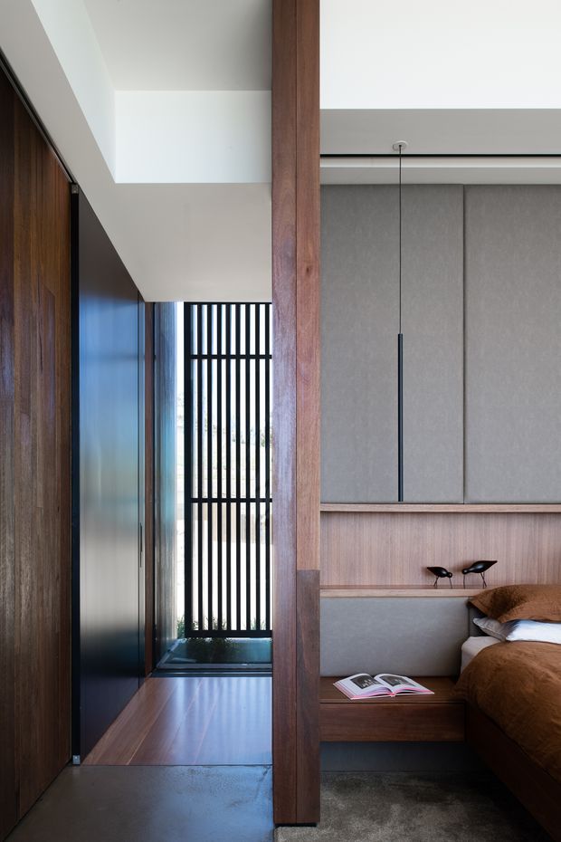 A refined contemporary bedroom interior viewed beside a narrow hallway or circulation space. The room features extensive dark timber built-in joinery, a substantial vertical timber post, a grey upholstered wall panel behind the bed, integrated timber bedside storage, and a slim black pendant light. A tall black vertical-slat screen brings filtered daylight into the passage, while polished dark flooring and restrained furnishings create a warm, minimalist atmosphere.