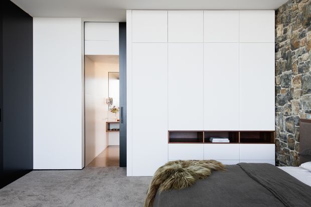 Modern bedroom with a low-profile bed in the foreground, dark grey bedding and a sheepskin throw. The room features extensive minimalist white built-in cabinetry with a dark open display niche, a deep grey carpeted floor, a dark painted wall at left, and a prominent rough natural stone feature wall at right. A doorway opens to a softly lit adjoining space.