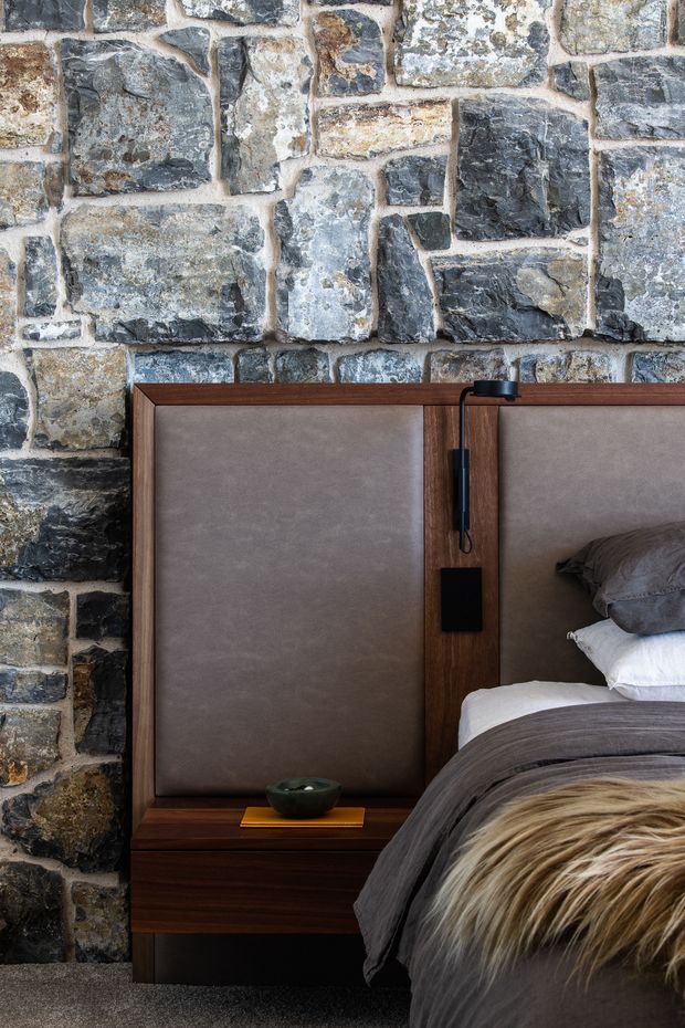 Close-up interior view of a bedroom focused on a sophisticated built-in timber bedhead and integrated bedside table. The bedhead has large taupe upholstered panels framed in dark timber, with a slim black reading light and a small black control panel. A rugged exposed natural stone wall forms the backdrop, while layered grey bedding, white pillows, and a textured fur throw are visible on the bed. The composition is warm, tactile, refined, and moody.
