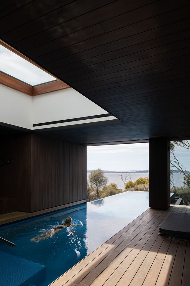A dramatic covered indoor-outdoor pool area with a dark timber-lined ceiling, timber wall panelling, and broad timber decking. An infinity-edge swimming pool extends toward a calm waterfront landscape visible through the open side. A skylight introduces daylight, while a dark built-in or upholstered poolside bench occupies the right foreground. The architecture has a strong contemporary, resort-like character with a seamless connection between interior and exterior.
