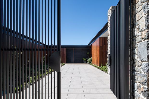 A contemporary residential entry driveway or forecourt viewed through an open black metal slatted gate. The space features pale large-format paved flooring, dark vertical screening, warm timber-clad walls, a dark garage or entry door, low planting, and a prominent rough natural-stone boundary wall. Strong linear elements and crisp detailing create a private, modern approach.