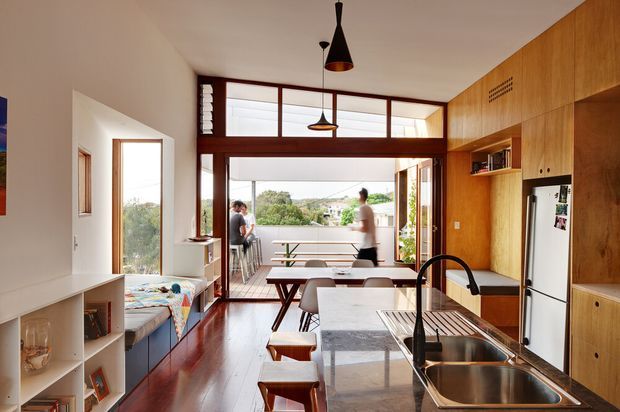 A warm, open-plan kitchen and dining area with a long stone island, double stainless-steel sink, timber cabinetry, integrated shelving, and a white refrigerator. Dark timber flooring and matching timber-framed glazing lead to a bright balcony or outdoor area beyond. A built-in window seat and dining table occupy the left side, while pendant lights hang over the kitchen and dining zones.