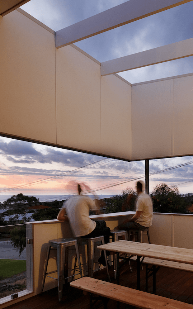 A covered rooftop or upper-level outdoor terrace overlooking a suburban coastal landscape at sunset. Two people sit at a narrow bar counter on metal stools beneath a translucent or open slatted canopy, with timber decking, a long timber picnic-style table and benches, and clear glass or transparent balustrading. The scene has a relaxed, informal hospitality-like atmosphere and broad sky views.