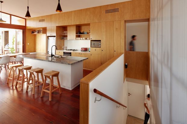 Warm open-plan kitchen and dining area with extensive light timber built-in cabinetry, a white island with a dark stone benchtop, timber stools, pendant lighting, and rich timber flooring. A stairwell with a timber handrail leads downward in the foreground, while glazed doors open to the exterior at left. The space has a clean, refined contemporary character with strong timber detailing.