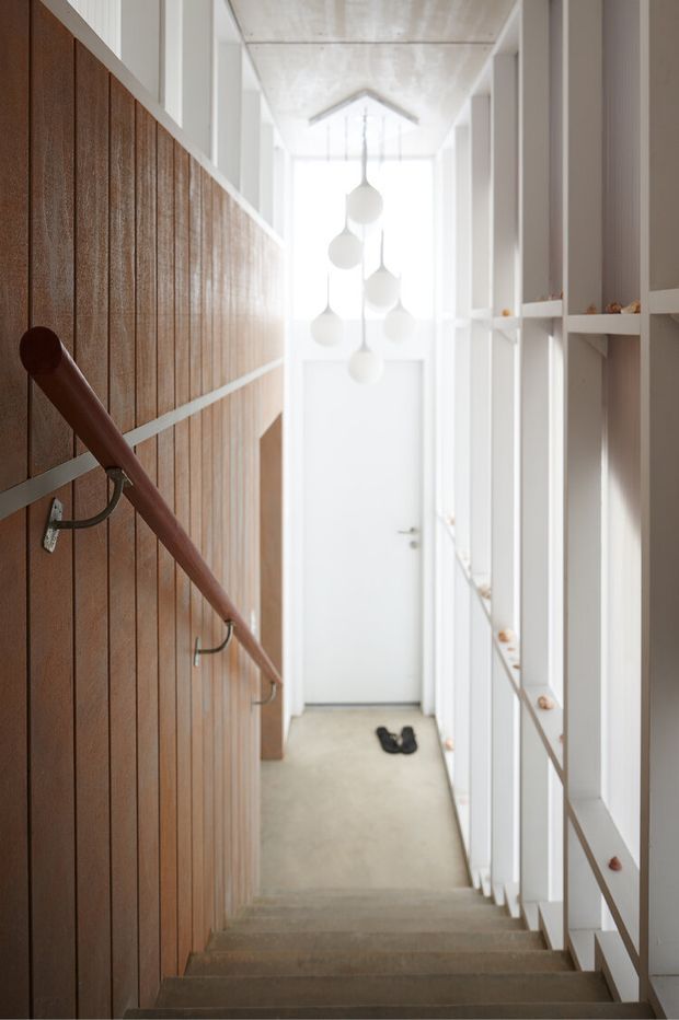 A narrow residential stairway viewed from above, with concrete steps, a warm vertical timber-paneled wall and timber handrail on the left, and white open shelving or balustrade elements on the right. A bright landing with a white door is visible at the bottom, illuminated by a cluster of sculptural white pendant lights. The design is minimalist and contemporary, with strong natural light and contrasting warm wood and white surfaces.