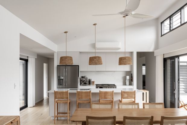 Bright open-plan kitchen and dining area with a white minimalist fit-out, light timber flooring, a central island with woven rattan bar stools, three woven pendant lights, stainless-steel appliances, and a timber dining table in the foreground. High windows and a ceiling fan contribute to an airy contemporary interior.