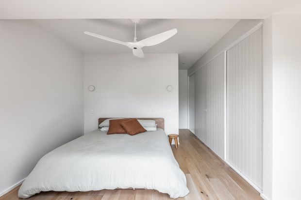 Bright, minimalist bedroom with a low-profile bed dressed in white bedding and warm brown cushions, set against white walls. A full-height bank of vertically slatted white wardrobe doors lines the right wall, while a light timber floor and sculptural white ceiling fan add subtle warmth and detail.