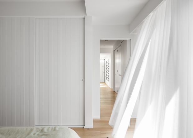Bright, minimalist bedroom view looking toward a long white hallway. The room features white vertical wall panelling or wardrobe doors, pale timber flooring, and sheer white curtains over a large window, with part of a bed visible in the foreground.