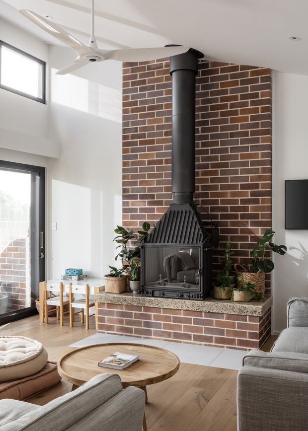 Bright contemporary living room with a prominent exposed red-brick fireplace chimney and black freestanding wood-burning stove. The room features pale timber flooring, light upholstered sofas, a round timber coffee table, indoor plants, large glazed doors and windows, and a white ceiling fan. A small dining area is visible beside the glazing.