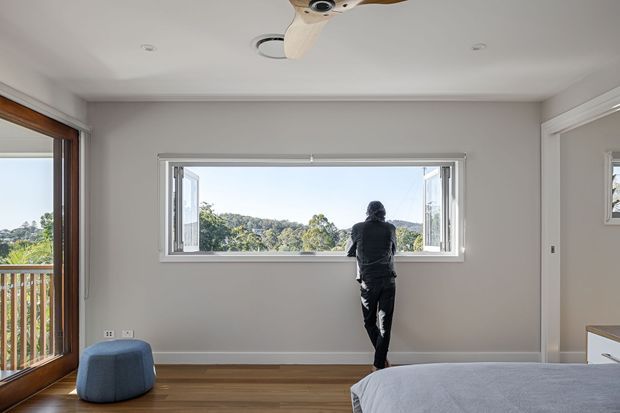 A bright, uncluttered bedroom with pale grey walls, timber flooring, a large open awning-style window overlooking a leafy suburban landscape, and a timber-framed glazed door opening to a balcony. A ceiling fan, small blue upholstered stool, and partial bed are visible. The room has a calm, contemporary residential character.