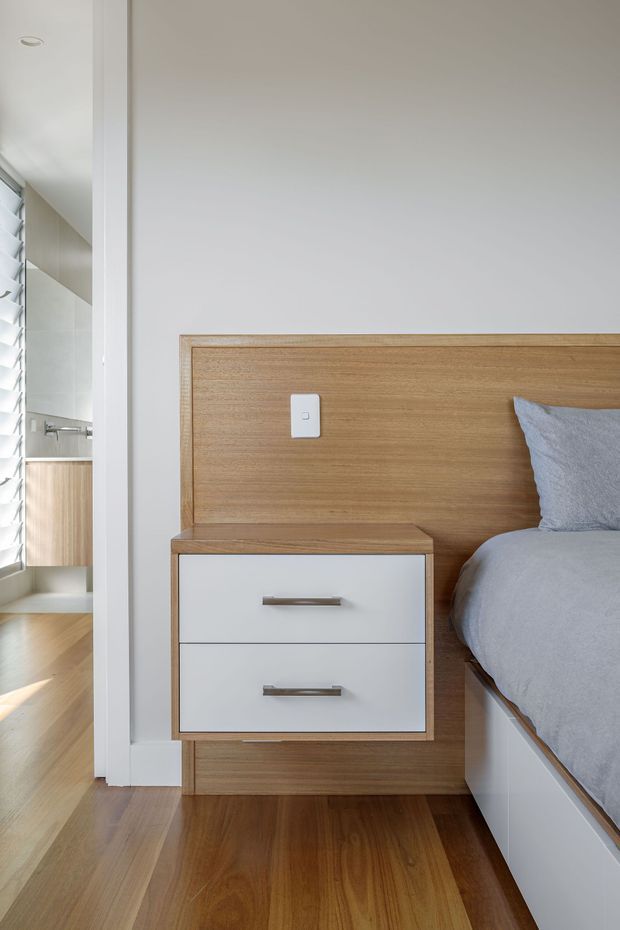 A bright, minimalist bedroom detail featuring a timber wall-mounted headboard integrated with a floating two-drawer bedside table. The bed has soft grey bedding and a grey upholstered pillow. Warm timber flooring continues through the room, with white walls, trim, and cabinetry. A louvred window and glimpse of an adjoining light-finished space are visible at left.
