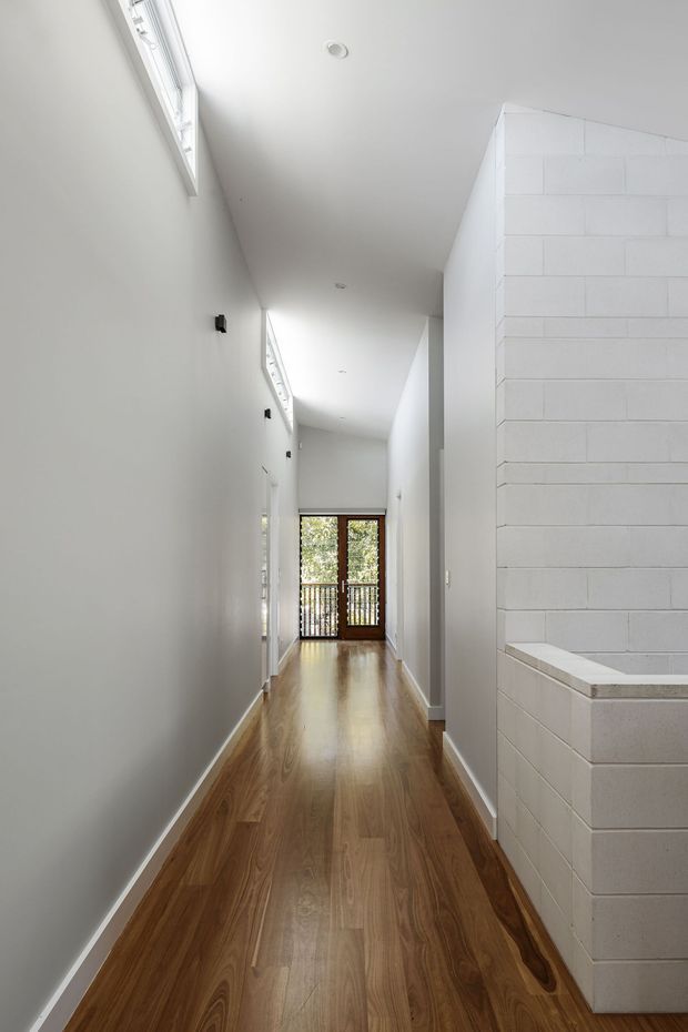 A long, narrow, bright residential hallway with polished warm timber flooring, smooth white walls and ceiling, high clerestory-style windows, recessed downlights, and a white painted brick feature wall enclosing a built-in structure on the right. Glazed timber-framed doors at the far end open toward an exterior balcony or landing.