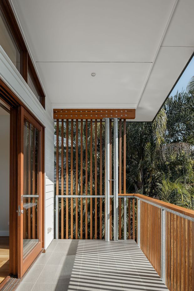 Covered outdoor balcony or terrace beside a timber-framed glazed door, with white weatherboard wall cladding, tiled flooring, timber and metal balustrades, and a vertical timber-and-metal privacy screen. Strong sunlight creates striped shadows across the floor, with tropical planting visible beyond the railing.