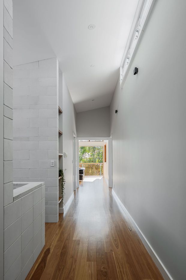 A bright, narrow interior hallway with warm timber flooring, white painted walls, and a high sloping ceiling. The left side features white tiled masonry and built-in open shelving, while a long high-level window brings in daylight along the right wall. The hallway leads to an open glazed doorway and timber balcony railing beyond. The space has a clean, restrained contemporary character.
