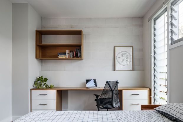 Bright, compact bedroom with a built-in home office along the wall. A timber wall-mounted bookshelf sits above a long desk with white drawer cabinetry, paired with a black ergonomic chair. The room has white painted masonry-style wall panels, pale walls, louvred windows providing abundant daylight, and a bed in the foreground with a geometric checked cover.