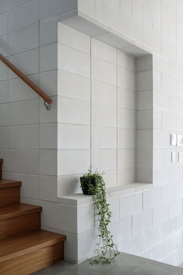 Interior architectural detail showing a stairway beside a deep recessed wall niche. The walls and niche are formed from painted white masonry blocks with visible horizontal and vertical joints. Warm timber stairs and a timber handrail contrast with the pale wall, while a trailing potted plant sits on the niche ledge. Electrical outlets and a small wall opening are visible on the right.