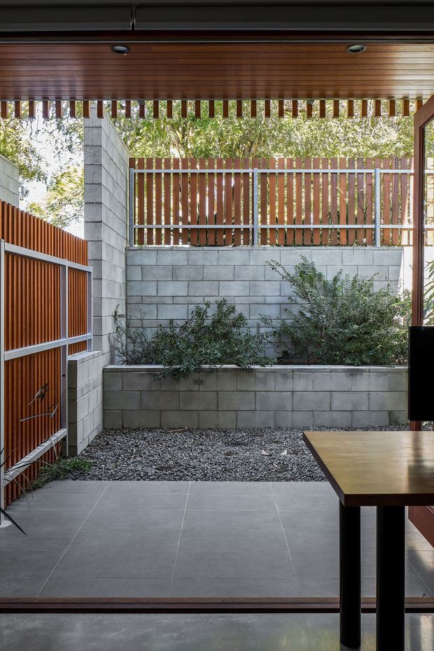 A covered semi-outdoor courtyard or patio viewed from inside, featuring grey tiled flooring, exposed concrete block walls, a raised planted bed, and vertical timber screening with metal framing. A timber-clad ceiling with recessed downlights provides shelter, while a partial dining table and chair are visible in the foreground. The space has a contemporary, restrained architectural character with strong indoor-outdoor connection.
