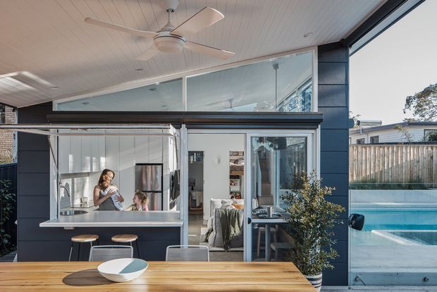 A bright contemporary indoor-outdoor living space with a compact kitchen opening onto a timber dining table and a swimming pool. The room features large glazed sliding doors, dark exterior wall cladding, a white-lined ceiling with a ceiling fan, stainless steel kitchen appliances, and a raised clerestory-style glazed section. A living area is visible beyond the kitchen, with outdoor fencing and pool glimpsed through the open glazing.