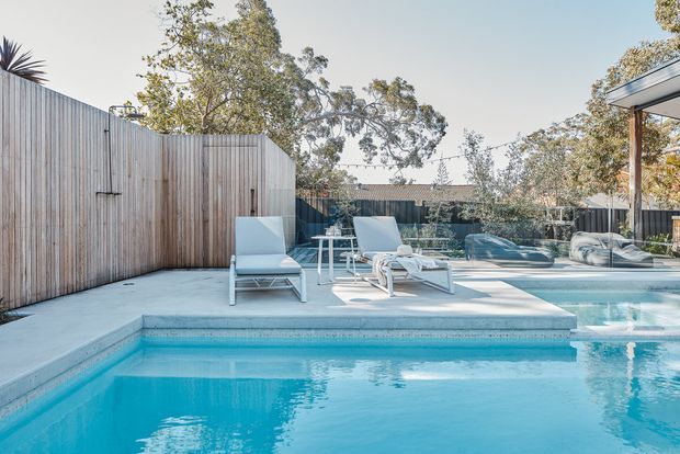 A bright contemporary outdoor pool area with a large turquoise swimming pool, pale concrete pool deck, white lounge chairs, and a covered outdoor seating area. Vertical timber privacy fencing encloses the space, with mature trees visible beyond. The scene has a clean, relaxed residential resort-like atmosphere.