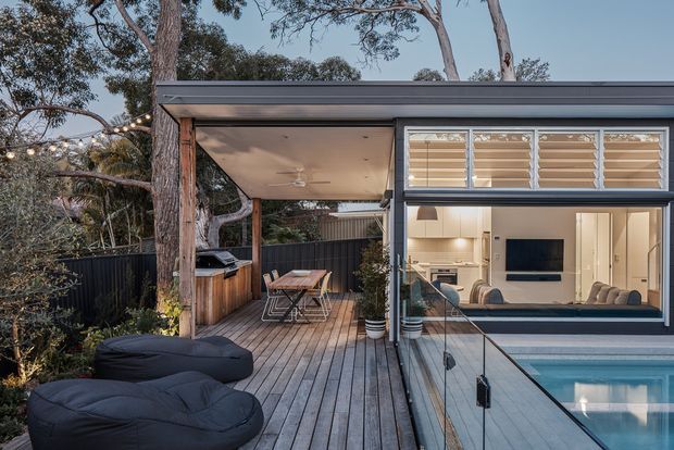 Contemporary indoor-outdoor living space with a timber deck, covered outdoor dining and barbecue area, and a swimming pool beside a glazed living room. The pavilion-like structure has a flat roof, extensive glazing, timber posts and decking, and louvred upper windows. Mature trees surround the deck, while the interior visible through the glazing includes a sofa, television, kitchen and pendant lighting.